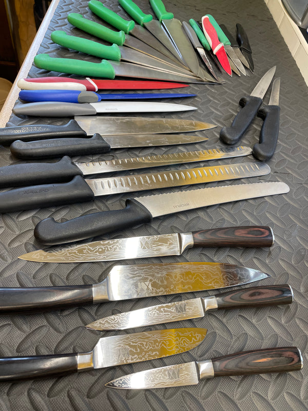 assorted kitchen knives laid out side by side on a textured rubber mat with a diamond plate pattern. The knives vary in size, blade shape, and handle design, including boning, fillet, carving, chef’s, cleaver, and santoku styles