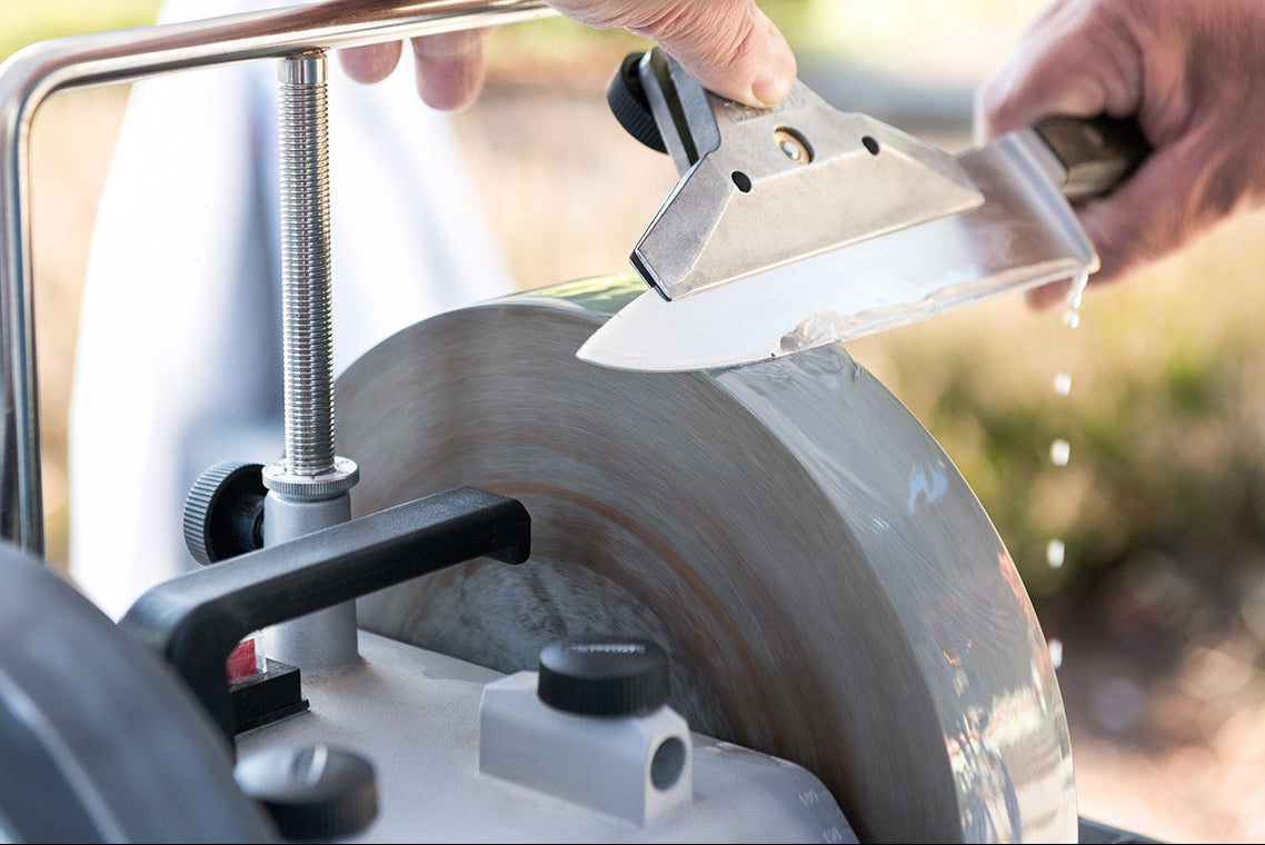 Person sharpening a large knife on a water-cooled grinding wheel at a professional sharpening station. The blade is held at a precise angle, with water dripping to cool the edge. The machine features a metal support bar and adjustment knobs for stability and control.
